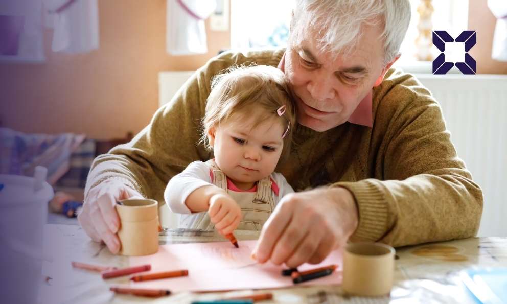 Grandfather guiding his granddaughter as they draw with crayons at a kitchen table.