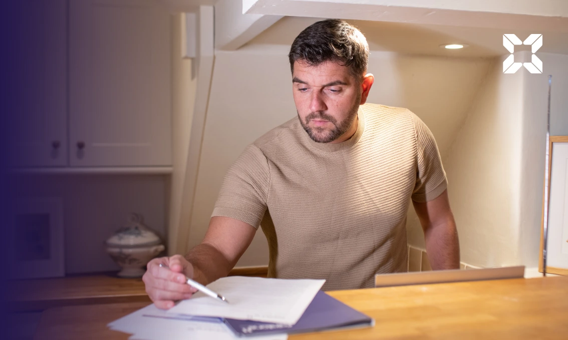 A man standing, looking at documents on a desk with a pen in his hand.