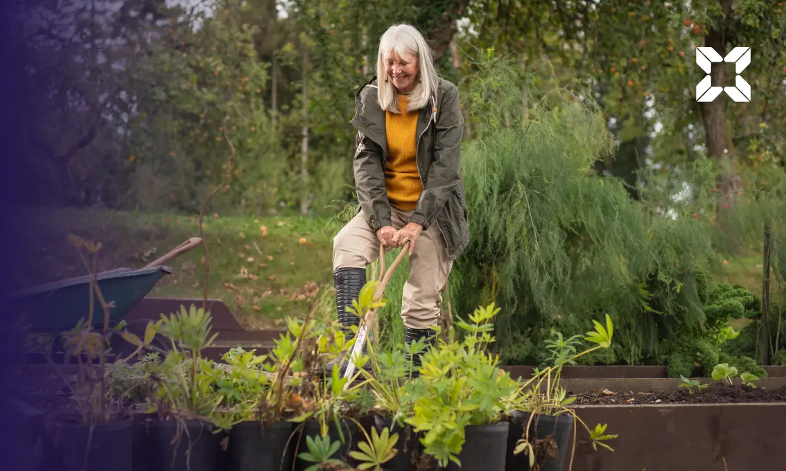 An elderly woman digging a vegetable patch holding a spade. She is smiling.