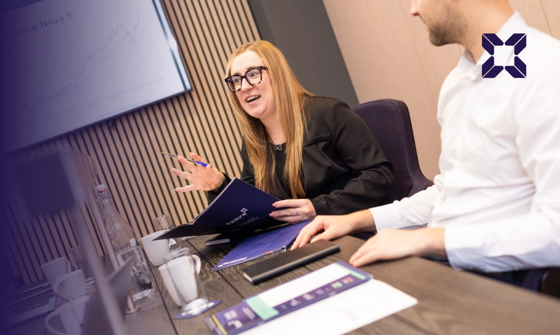 A lady with glasses is sat at a table explaining something to her colleagues. She has a screen behind her.