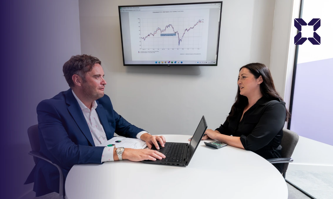 A man and a woman sat opposite each other in an office across a desk, chatting. There is an image of a graph displayed on a monitor on the wall.