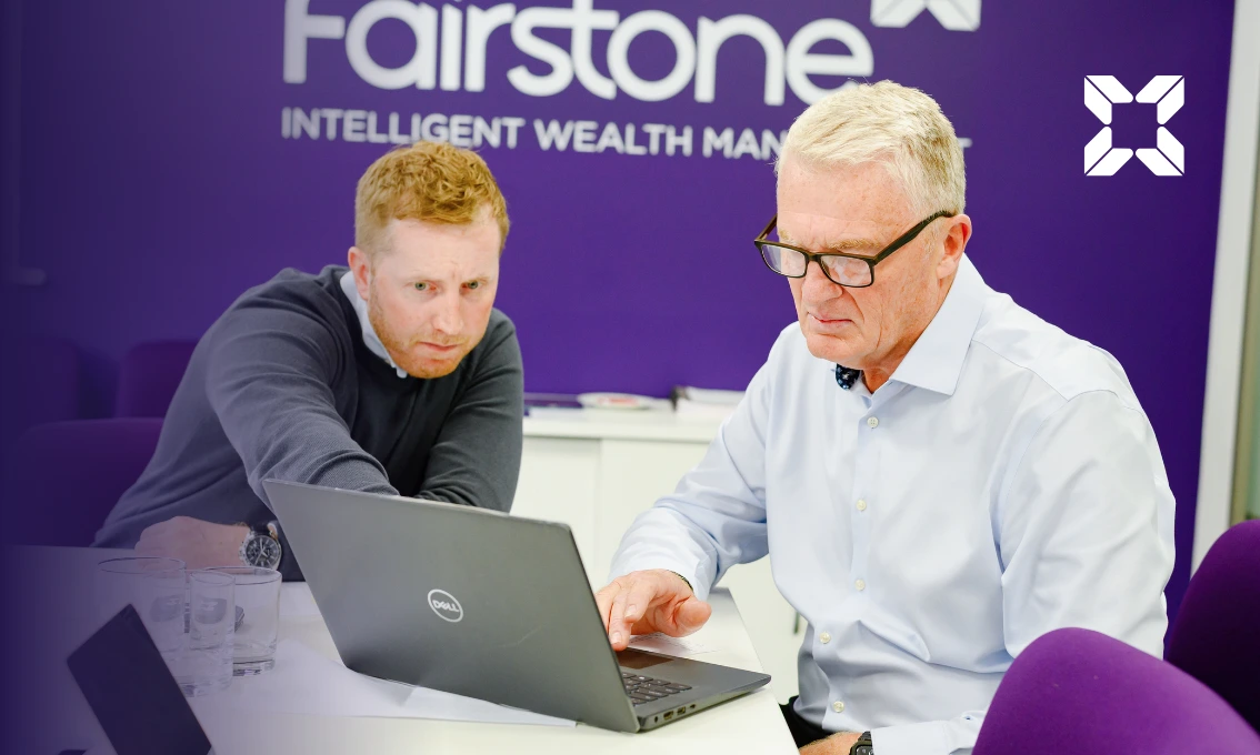 A younger man and an older man are sat at a desk looking at a laptop