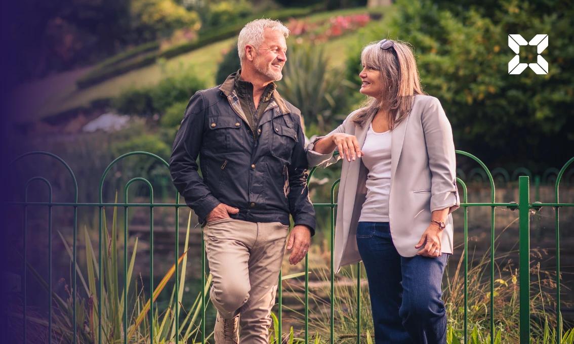 An older male and female couple leaning against a railing in a park smiling at each other.