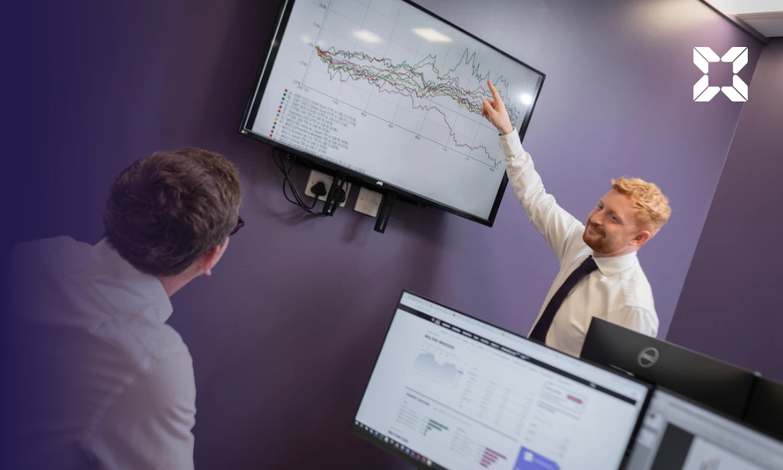 A man is standing pointing to a monitor in an office showing line graphs. He is talking to a colleague who is sat down.