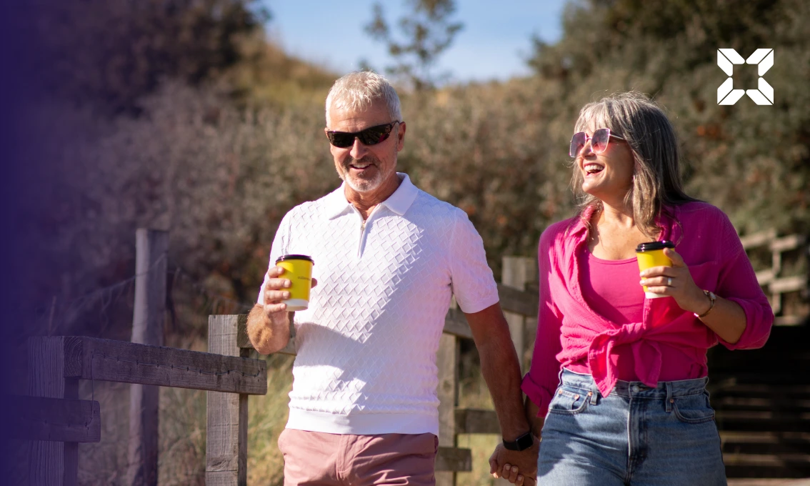 A middle aged male and female couple are walking down some steps towards a beach. It is sunny, they are each carrying a coffee and are smiling at each other.