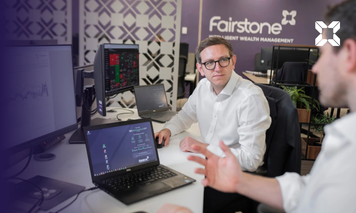 Two male colleagues are sat at a desk having a conversation with laptops in front of them.